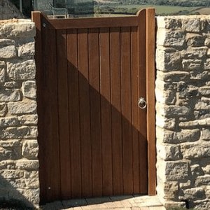 A hardwood gate with a rich brown finish is set between two stone walls, featuring vertical wooden planks and a silver ring handle. Sunlight casts a diagonal shadow on the gate, highlighting its sturdy construction.