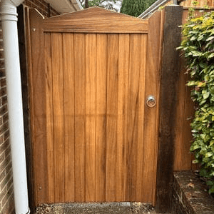 A sturdy hardwood gate with a vertical plank design stands between a brick wall and a wooden fence. The warm, natural wood tones complement the outdoor setting, while the curved top adds a touch of elegance. A silver ring handle provides secure access, and the surrounding greenery enhances the gate’s rustic charm.